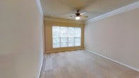 This single-floor space features a well-proportioned room finished with warm, neutral-toned walls and plush wall-to-wall carpeting that adds comfort underfoot. Crown molding runs along the perimeter where the walls meet the textured ceiling, lending a clean, classic architectural detail to the interior. A ceiling fan with an integrated light fixture is centrally positioned, providing both air circulation and ambient lighting throughout the room. A large, multi-pane window dominates one wall, allowing generous natural light to filter into the space while offering views of the surrounding greenery outside. The window's bright exposure contributes to an open, airy atmosphere. White baseboards provide a crisp contrast against the neutral walls and carpet, maintaining a cohesive and tidy aesthetic. The room's layout is open and versatile, offering flexibility in how the space can be arranged or utilized. Overall, the room presents a straightforward, comfortable environment characterized by practical features and a calm, neutral palette suited to a variety of uses.