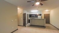 This single-floor space offers an open-concept layout that seamlessly connects the living area to the kitchen. The living area features plush carpeting and a ceiling fan with an integrated light fixture, providing both comfort and functionality. Neutral-toned walls with dark baseboards create a clean, modern aesthetic throughout.

The kitchen is well-appointed with dark cabinetry, a stainless steel refrigerator, and a built-in microwave. A peninsula with a light-toned countertop serves as a natural divider between the kitchen and living space, offering additional prep space and seating potential. Pendant lighting above the peninsula adds a stylish focal point, complemented by recessed lighting overhead.

Multiple doorways visible from the main area suggest additional rooms branching off the central space, providing a functional flow throughout the unit. The combination of carpet in the living zone and hard flooring in the kitchen area adds practical distinction between spaces. Overall, the unit presents a contemporary and well-organized layout suited for everyday living.