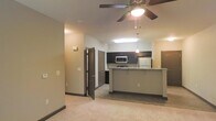 This single-floor space offers an open-concept layout that seamlessly connects the living area to the kitchen. The living area features plush carpeting and a ceiling fan with an integrated light fixture, providing both comfort and functionality. Neutral-toned walls with dark baseboards create a clean, modern aesthetic throughout.

The kitchen is well-appointed with dark cabinetry, a stainless steel refrigerator, and a built-in microwave. A peninsula with a light-toned countertop serves as a natural divider between the kitchen and living space, offering additional prep space and seating potential. Pendant lighting above the peninsula adds a stylish focal point, complemented by recessed lighting overhead.

Multiple doorways visible from the main area suggest additional rooms branching off the central space, providing a functional flow throughout the unit. The combination of carpet in the living zone and hard flooring in the kitchen area adds practical distinction between spaces. Overall, the unit presents a contemporary and well-organized layout suited for everyday living.