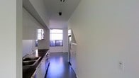 This single-floor space showcases a clean, contemporary design aesthetic defined by a minimalist sensibility and modern finishes throughout. The kitchen is a standout feature, laid out in a galley-style configuration that maximizes efficiency. It is equipped with stainless steel appliances, including a full-sized range with multiple burners and a large refrigerator, complemented by dark countertops that contrast sharply against crisp white cabinetry. The deep, dark hardwood flooring runs the length of the space, adding warmth and a polished look underfoot. High ceilings amplify the sense of openness, while large windows along the far wall invite generous amounts of natural light, creating a bright and airy atmosphere. The windows feature horizontal blinds, offering adjustable light control and privacy. Recessed and track lighting fixtures mounted on the ceiling provide additional illumination, ensuring the space is well-lit at all times. The walls are finished in a clean, neutral white, reinforcing the modern and uncluttered character of the space. Overall, the design balances functionality with a refined aesthetic, making it a well-appointed and practical living environment.