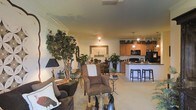 This single-floor residence showcases an open-concept layout that integrates the living, dining, and kitchen areas into one cohesive, flowing space. The living area is generously sized, anchored by plush, upholstered seating and accented by large decorative indoor trees that add a natural, organic element to the interior. Crown molding runs along the ceiling, adding a refined architectural detail to the space.

The dining area transitions naturally from the living zone, furnished with a dark wood table and cushioned chairs, creating an intimate setting for meals. A framed botanical artwork adorns the wall nearby, contributing to the warm, nature-inspired aesthetic carried throughout.

The kitchen is well-appointed with warm wood cabinetry, dark appliances including a refrigerator and range, and a peninsula with bar-height seating for two. Pendant lighting illuminates the counter space, which features granite-style surfaces. Upper cabinetry extends to the ceiling, maximizing storage.

Neutral carpeting flows throughout the main living spaces, complementing the warm tones of the furnishings and cabinetry. Overall, the space offers a comfortable, well-organized layout with a cohesive and inviting ambiance.