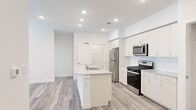 This single-floor space showcases a clean, contemporary design defined by bright white walls, light wood-tone flooring, and recessed lighting throughout. The open-concept kitchen is a standout feature, equipped with stainless steel appliances including a gas range, over-the-range microwave, and refrigerator. White shaker-style cabinetry extends to the upper walls, complemented by granite countertops. A central island with an integrated sink and additional granite surface provides both workspace and a natural dividing point between the kitchen and the open living area beyond. Adjacent to the kitchen, a built-in desk nook with open shelving and granite countertop offers a dedicated workspace. The living area benefits from natural light through large windows fitted with blinds and is outfitted with a ceiling fan. A dedicated laundry area houses a full-sized washer and dryer. Storage is well-addressed with a spacious walk-in closet featuring wire shelving on multiple walls. The consistent flooring, neutral palette, and modern finishes create a cohesive, functional ambiance throughout the entire space.