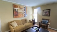 This single-floor space offers a comfortable and well-appointed living area that exudes a classic, homey charm. The living room is anchored by a plush, tan sofa adorned with decorative pillows, complemented by a patterned armchair that adds a touch of traditional elegance. Crown molding traces the ceiling perimeter, lending the room a refined architectural detail. A large, bold abstract painting in warm earth tones — featuring shades of brown, rust, and blue — serves as a striking focal point on one wall, adding a contemporary artistic flair to the otherwise classic setting. Natural light filters in through a window dressed with horizontal blinds, brightening the space and creating a pleasant ambiance. A dark circular side table with a table lamp provides functional accent lighting, while hardwood flooring anchors the room with warmth and durability. A dark coffee table with a tray, wine bottle, and glassware adds an inviting, lived-in quality. A framed picture on the adjacent wall completes the decor. Overall, this space presents a cozy and tastefully decorated environment well-suited for relaxed everyday living.