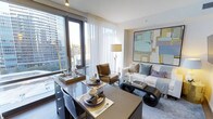 This single-floor residence showcases a seamlessly integrated open-plan layout where the dining and living areas share a bright, cohesive space. Floor-to-ceiling windows dominate one wall, flooding the interior with natural light and framing impressive elevated views of the surrounding urban skyline and greenery below. Sheer white curtains complement the windows, softening the light while maintaining an airy atmosphere.

The dining area features a sleek, dark-toned table set with elegant dishware, gold-accented placemats, and neatly folded napkins, conveying a refined, sophisticated aesthetic. Adjacent to it, the living area is anchored by a cream sofa adorned with blue and grey accent cushions, accompanied by a cognac leather accent chair and a sculptural dark coffee table. A large abstract artwork in muted blues, yellows, and purples serves as a focal point on the wall.

Throughout, the design palette balances neutral tones with warm metallic accents, creating an upscale yet inviting ambiance. Clean lines, high-quality furnishings, and thoughtful styling give the space a polished, contemporary character well-suited to urban living.