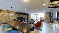 Located at 1516 Xavier Street in Denver's vibrant 80204 corridor, this single-floor communal space is thoughtfully designed to encourage both collaboration and relaxation. The open-plan layout features a variety of distinct seating zones, seamlessly connected by polished tile flooring and a clean, contemporary aesthetic. A dramatic stacked stone accent wall serves as the architectural centerpiece, adding warmth and texture to the otherwise modern interior. Large floor-to-ceiling windows line one side of the space, flooding the interior with natural light and offering views of the surrounding cityscape. A long, dark-wood communal table with colorful bar-height chairs in blue and green provides an ideal setting for group work or casual gatherings. Adjacent lounge areas are furnished with plush sofas, armchairs in bold orange and blue, and low coffee tables, creating a relaxed atmosphere suited for informal meetings or quiet downtime. Pendant lighting with woven shades and a black grid ceiling fixture add stylish layers of illumination. Private high-back booth seating near the accent wall offers a more secluded retreat within the open environment. Access to an outdoor area is available through glass doors, extending the usable space beyond the interior. Overall, the space balances modern design with inviting comfort.