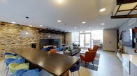 Located at 1516 Xavier Street in Denver's vibrant 80204 corridor, this single-floor communal space is thoughtfully designed to encourage both collaboration and relaxation. The open-plan layout features a variety of distinct seating zones, seamlessly connected by polished tile flooring and a clean, contemporary aesthetic. A dramatic stacked stone accent wall serves as the architectural centerpiece, adding warmth and texture to the otherwise modern interior. Large floor-to-ceiling windows line one side of the space, flooding the interior with natural light and offering views of the surrounding cityscape. A long, dark-wood communal table with colorful bar-height chairs in blue and green provides an ideal setting for group work or casual gatherings. Adjacent lounge areas are furnished with plush sofas, armchairs in bold orange and blue, and low coffee tables, creating a relaxed atmosphere suited for informal meetings or quiet downtime. Pendant lighting with woven shades and a black grid ceiling fixture add stylish layers of illumination. Private high-back booth seating near the accent wall offers a more secluded retreat within the open environment. Access to an outdoor area is available through glass doors, extending the usable space beyond the interior. Overall, the space balances modern design with inviting comfort.