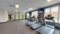This contemporary fitness center is thoughtfully designed to accommodate a wide range of workout styles and fitness goals. The facility is organized into distinct zones, each tailored to specific training needs. The cardio area features a row of modern treadmills and elliptical machines, all positioned facing large windows that bring in natural light and offer views of the surrounding exterior. Wall-mounted televisions are conveniently placed throughout for entertainment during workouts. The strength training section is well-stocked with a full dumbbell rack backed by floor-to-ceiling mirrors, adjustable benches, a power rack, cable machines, and a functional trainer, providing ample options for resistance-based exercise. Plyo boxes and additional cable equipment occupy a dedicated functional training zone, separated from the main floor by sleek glass partition walls with dark metal framing. A spacious studio room with light wood-look flooring, TRX suspension straps, wall-mounted bar ladders, and a large display screen offers an ideal environment for group fitness classes, yoga, or personal training sessions. Throughout the facility, durable rubber flooring, recessed and panel lighting, and a clean neutral color palette contribute to a professional and energizing atmosphere.