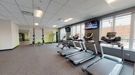 This contemporary fitness center is thoughtfully designed to accommodate a wide range of workout styles and fitness goals. The facility is organized into distinct zones, each tailored to specific training needs. The cardio area features a row of modern treadmills and elliptical machines, all positioned facing large windows that bring in natural light and offer views of the surrounding exterior. Wall-mounted televisions are conveniently placed throughout for entertainment during workouts. The strength training section is well-stocked with a full dumbbell rack backed by floor-to-ceiling mirrors, adjustable benches, a power rack, cable machines, and a functional trainer, providing ample options for resistance-based exercise. Plyo boxes and additional cable equipment occupy a dedicated functional training zone, separated from the main floor by sleek glass partition walls with dark metal framing. A spacious studio room with light wood-look flooring, TRX suspension straps, wall-mounted bar ladders, and a large display screen offers an ideal environment for group fitness classes, yoga, or personal training sessions. Throughout the facility, durable rubber flooring, recessed and panel lighting, and a clean neutral color palette contribute to a professional and energizing atmosphere.