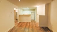 This single-floor space is designed with an open, functional layout that maximizes the sense of roominess. The main living area flows seamlessly toward the kitchen, which is defined by a peninsula-style breakfast bar that serves as a natural divider without enclosing the space. The kitchen is finished with white cabinetry, a white refrigerator, and overhead fluorescent lighting that brightens the cooking area. White walls and white trim throughout contribute to a clean, neutral aesthetic.

Warm-toned hardwood flooring runs across the main living space, adding character and a sense of continuity between areas. Recessed lighting in the living area complements the natural brightness of the space. A hallway visible to the left leads to additional rooms, with a glimpse of what appears to be a bathroom featuring a vanity mirror. To the right of the kitchen, a partial wall with an electrical panel is visible, indicating a practical, utility-conscious layout.

The overall ambiance is bright, clean, and move-in ready, with a straightforward design that prioritizes open sightlines and ease of movement throughout the single-floor layout.