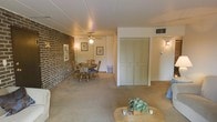 This single-floor space offers an open, combined living and dining layout that feels welcoming and functional. A striking full-height exposed brick wall runs along one side of the room, adding a rustic, textural contrast to the otherwise neutral interior. The ceiling is flat and features a central ceiling fan with a light fixture, providing both air circulation and ambient lighting, while wall-mounted sconces add supplemental warmth throughout the space.

The dining area is positioned near the brick wall and accommodates a wooden table with chairs featuring blue upholstered seats, creating a casual and comfortable setting. Adjacent to the dining area, a set of double louvered closet doors in a muted sage green offers generous built-in storage, a practical feature that enhances the room's organization.

The flooring throughout is carpeted in a neutral beige tone, contributing to a soft, cozy ambiance. The living area is defined by a wicker coffee table adorned with a small plant arrangement. A ceiling vent is visible, indicating a central HVAC system. Framed artwork on the walls adds a personal, lived-in character. Overall, the space presents a functional and modest layout with warm, approachable design details.