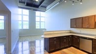 This single-floor space showcases an open, loft-inspired layout that blends industrial and contemporary design elements. The striking exposed ceiling features dark-painted wood beams and visible ductwork, complemented by a ceiling fan and adjustable track lighting that provide both function and visual interest. Large, multi-pane windows flood the interior with abundant natural light and offer views of surrounding greenery, creating a bright and airy atmosphere throughout.

The hardwood flooring, with its warm tones and polished finish, adds a refined quality to the open floor plan. A substantial L-shaped cabinetry unit serves as a central feature, finished in rich dark wood with brushed nickel hardware and topped with a granite or stone-look countertop. The unit provides generous storage and workspace, doubling as a natural divider within the open layout. Upper cabinetry on the adjacent wall adds additional storage. The generously proportioned, light-filled space and thoughtful combination of industrial and refined finishes give this property a versatile, modern character suitable for a variety of uses.