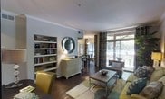 This single-floor space showcases a thoughtfully designed open-concept layout that seamlessly connects the living and dining areas. The living area is anchored by a neutral area rug and features a contemporary coffee table with a chrome base, complemented by teal and yellow accent chairs that introduce a vibrant pop of color. A built-in bookcase lines one wall, offering both functional storage and a decorative display area for books and personal items. A decorative round mirror with a mosaic frame serves as a striking focal point on the wall above a white chest of drawers, adding visual depth to the space. Rich, dark hardwood flooring runs throughout, providing warmth and contrast against the light gray walls and white crown molding. Floor-to-ceiling windows dressed with patterned curtains flood the interior with natural light while offering views of an outdoor seating area beyond. The dining area is visible in the background, furnished with bar-height seating and a pendant light fixture, creating a distinct yet connected zone within the open layout.
