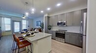 This efficiently designed 1-bedroom, 1-bathroom residence offers a thoughtfully planned layout across a single floor. The open-concept kitchen and living area serves as the heart of the unit, where sleek gray cabinetry with brushed silver hardware lines the kitchen walls, complemented by a classic white subway tile backsplash, granite countertops, and a full suite of stainless steel appliances including a range, over-the-range microwave, and refrigerator. A central kitchen island with a granite countertop and built-in sink provides additional prep space and doubles as a breakfast bar with room for seating. Stylish glass pendant lights hang above the island, adding a modern touch. Light wood-style flooring runs throughout, enhancing the bright and airy feel of the space. The adjoining living area offers a comfortable, open setting with natural light filtering in through windows. The bedroom is a well-proportioned private retreat featuring a ceiling fan, a window with natural light, and direct access to the bathroom. The bathroom continues the cohesive design language with gray cabinetry, granite countertops, a large mirror, and a tiled soaking tub and shower combination, finished with marble-look tile surround.
