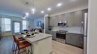 This efficiently designed 1-bedroom, 1-bathroom residence offers a thoughtfully planned layout across a single floor. The open-concept kitchen and living area serves as the heart of the unit, where sleek gray cabinetry with brushed silver hardware lines the kitchen walls, complemented by a classic white subway tile backsplash, granite countertops, and a full suite of stainless steel appliances including a range, over-the-range microwave, and refrigerator. A central kitchen island with a granite countertop and built-in sink provides additional prep space and doubles as a breakfast bar with room for seating. Stylish glass pendant lights hang above the island, adding a modern touch. Light wood-style flooring runs throughout, enhancing the bright and airy feel of the space. The adjoining living area offers a comfortable, open setting with natural light filtering in through windows. The bedroom is a well-proportioned private retreat featuring a ceiling fan, a window with natural light, and direct access to the bathroom. The bathroom continues the cohesive design language with gray cabinetry, granite countertops, a large mirror, and a tiled soaking tub and shower combination, finished with marble-look tile surround.