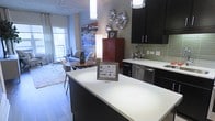 This single-floor space showcases a thoughtfully designed open-concept layout that seamlessly connects the kitchen, dining, and living areas. The kitchen is a standout feature, equipped with sleek dark cabinetry that extends to the upper walls, paired with crisp white countertops that provide a striking contrast. A generously sized central island serves as both a functional workspace and a visual anchor for the room. Green glass tile backsplash adds a refined touch of color, while stainless steel appliances, including a dishwasher, contribute to a polished, contemporary aesthetic. Pendant lighting above the island and a tall floor lamp in the living area provide layered illumination throughout the space. The dining area accommodates a small round table with seating, transitioning naturally into the living area, which features a comfortable seating arrangement. Light hardwood flooring runs throughout, enhancing the sense of openness. A large window floods the living area with natural light, adding warmth to the overall cool-toned palette of blues and grays. Decorative wall art and sculptural accents add personality and visual interest throughout.