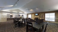This single-floor communal space is thoughtfully designed to accommodate both dining and lounging in a warm, inviting atmosphere. The layout is open and expansive, seamlessly connecting multiple functional zones within one cohesive room.

The dining area features several dark-stained tables and matching chairs arranged to encourage group gatherings, complemented by track lighting overhead that provides ample illumination throughout the space. Vaulted ceilings with angled planes add architectural interest and a sense of openness, while neutral wall tones and decorative floral artwork contribute to the welcoming ambiance.

Transitioning into the lounge area, a classic fireplace serves as a natural focal point, framed by a clean white mantel and accented by a wall-mounted flat-screen television above. Plush, dark upholstered armchairs are arranged nearby, creating a comfortable seating nook ideal for relaxation or casual conversation. Built-in shelving units on either side of this area provide display space and add to the room's refined character.

Natural light filters in through multiple windows, brightening the interior alongside the track and ceiling lighting. Dark hardwood-style flooring runs throughout, grounding the space and tying together the dining and lounge areas cohesively. Overall, this space strikes a balance between functionality and comfort.