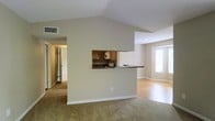 This single-floor residence showcases a thoughtfully designed open-concept layout that seamlessly connects the living and dining areas. A striking vaulted ceiling rises above the main living space, adding a sense of vertical volume and airiness to the interior. The living area is finished with plush wall-to-wall carpeting, while an adjacent space transitions to warm hardwood flooring, providing a clear visual distinction between zones.

A central dividing wall features a pass-through kitchen window, complete with a built-in countertop overhang that functions as a breakfast bar. The kitchen, visible through the opening, includes medium-toned wood cabinetry and under-cabinet appliances, offering practical storage and workspace. The pass-through design maintains an open, connected feel between the kitchen and main living areas.

Natural light filters into the space through a window in the adjoining room, brightening the neutral beige walls and white trim throughout. A hallway visible on the left side of the space leads to additional rooms, suggesting a functional and well-organized floor plan. The overall aesthetic is clean, neutral, and versatile.