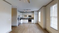 This single-floor space offers a clean, modern aesthetic defined by an open-concept layout that seamlessly connects the kitchen and living areas. The kitchen is a standout feature, showcasing crisp white shaker-style cabinetry paired with dark countertops for a striking contrast. Stainless steel appliances, including a French door refrigerator with ice and water dispenser, a range, and an over-the-range microwave, provide a fully equipped cooking environment. Track lighting above the kitchen workspace ensures excellent task illumination, while a ceiling fan in the adjoining living area promotes comfortable airflow throughout. Light wood-tone luxury vinyl plank flooring runs throughout the main living space, lending warmth and continuity to the interior. Neutral-toned walls and white trim create a bright, inviting atmosphere, complemented by natural light streaming in through a window fitted with white blinds. Crown molding adds a refined architectural detail to the transition between rooms. The layout is practical and well-organized, offering a welcoming, move-in-ready environment with thoughtful design details that balance functionality and style.