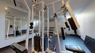 Located at 1551 Riverside Dr in Ottawa, Ontario, this dedicated exercise room offers a well-equipped fitness space designed to support a variety of strength training routines. The room features durable speckled rubber flooring throughout, providing both safety and resilience underfoot. A standout feature is the large wall-mounted mirror along one side, which visually expands the space and allows users to monitor their form during workouts.

The room is outfitted with a multi-station cable machine capable of supporting a wide range of resistance exercises, complemented by a flat weight bench and a barbell rack with weight plates for free weight training. A padded floor mat is available for stretching or floor-based exercises. Overhead recessed lighting, combined with a window that allows natural light to filter in, keeps the space bright and well-lit. A wall-mounted air conditioning unit ensures year-round climate comfort.

The white walls and light ceiling create a clean, functional aesthetic, while a security camera provides an added sense of safety. The overall layout is efficiently organized, making practical use of the available floor area to accommodate multiple pieces of equipment without feeling cramped. This exercise room serves as a practical and well-maintained amenity for residents.