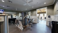 This fitness center is thoughtfully designed across two interconnected zones, offering a comprehensive workout environment for a variety of exercise routines. The strength training area features dark olive-green walls accented by white wainscoting, crown molding, and large wall-mounted mirrors that amplify the sense of space. This zone is outfitted with a multi-station cable machine, a rack of dumbbells, a barbell squat rack with bench, and a wall-mounted television, all set on durable rubber flooring. A ceiling fan and recessed lighting ensure comfort during intense sessions.

The adjacent cardio zone is architecturally distinct, showcasing a vaulted ceiling finished in warm natural wood with exposed dark beams, lending the space a bright, airy character. A row of large windows lines the walls, flooding the room with natural light and offering outdoor views. TRUE-brand treadmills, elliptical trainers, upright bikes, and recumbent bikes are neatly arranged throughout. A dedicated functional fitness nook features a built-in arched alcove stocked with stability balls, resistance bands, medicine ball racks, plyo boxes, and yoga mats — providing ample resources for dynamic and restorative training alike.