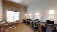 This business center is a thoughtfully designed, single-floor workspace that balances functionality with a clean, modern aesthetic. The main workstation area features a continuous run of dark wood cabinetry-style desks lining the walls, accommodating multiple computer terminals separated by frosted glass privacy dividers. Pendant lighting hangs from the notably tall ceilings, casting focused illumination over each workstation, while ergonomic task chairs provide comfortable seating for extended work sessions. A printer and additional office equipment are readily accessible along the shared desk surface.

Natural light enters through windows dressed with layered blinds and sheer curtains, contributing to a bright yet controlled atmosphere. A secondary area within the space offers a round bistro-style table paired with slatted wood chairs, creating an informal setting for small meetings or casual work. Nearby, a wood credenza displays brochures and informational materials beneath a trio of framed artwork, adding a polished, welcoming touch. Patterned carpeting throughout softens the space acoustically and visually, while neutral wall tones and warm wood finishes maintain a cohesive, professional ambiance throughout.