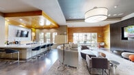 Located at 1550 Crescent Pointe Pkwy in College Station, Texas, this single-floor community clubhouse offers a generously sized, open-plan gathering space designed with a modern, upscale aesthetic. The entrance foyer welcomes visitors with polished concrete floors, high ceilings, and glass-paneled double doors that frame views into the main lounge beyond, with colorful modular seating providing an inviting first impression.

The main lounge area is anchored by a large drum pendant light and features a tray ceiling with recessed lighting. Warm natural wood wall and ceiling panels contrast beautifully with dark accent walls, one of which hosts a flat-screen television. Plush curved sofas, contemporary lounge chairs, and a textured area rug define a comfortable seating zone, while bold abstract artwork adds vibrant pops of color throughout.

An open kitchenette and bar area sits adjacent to the lounge, complete with bar-height seating, pendant lighting, and tile backsplash details beneath a warm wood ceiling canopy. A casual dining nook with a round orange-accented table and modern chairs sits nearby.

The game room zone features polished concrete flooring, a full-size pool table, a shuffleboard table, and expansive windows that flood the space with natural light and offer outdoor views of the surrounding property. Together, these areas create a dynamic, well-appointed communal space ideal for socializing and recreation.