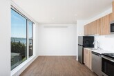This single-floor unit showcases a clean, modern design throughout, combining light wood-tone finishes with crisp white walls and thoughtful layout. The open living area is a standout feature, anchored by expansive floor-to-ceiling windows that flood the space with natural light and frame scenic views of the surrounding treetops and neighborhood rooftops. Light hardwood-style flooring extends across the main living area, adding warmth and continuity.

The kitchen is efficiently designed with natural wood-grain cabinetry extending to the ceiling, providing ample storage. A full suite of stainless steel appliances — including a range, over-the-range microwave, dishwasher, and refrigerator — is complemented by clean white countertops and a sleek single-basin sink with a modern faucet. A subtle backsplash adds a refined touch.

The bedroom offers a quieter, more intimate setting with soft carpeted flooring, white walls, a closet, and a window with a blind for privacy and natural light control. A wall-mounted HVAC unit provides climate control.

The bathroom is finished in a clean, minimalist style, featuring a wood-tone vanity with a white countertop and under-mount sink, a wall-mounted light fixture, a large mirror, and a walk-in shower with chrome fixtures and a towel bar. The overall ambiance of the unit is bright, fresh, and contemporary.