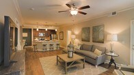 This single-floor residence showcases an open-concept layout that seamlessly integrates the living area and kitchen into one cohesive, inviting space. The living area is anchored by a ceiling fan with a light fixture, crown molding, and warm neutral-toned walls that contribute to a comfortable, welcoming ambiance. Large artwork depicting natural tree silhouettes decorates the walls, enhancing the space's understated aesthetic. The living area flows naturally into the kitchen, which features rich dark wood cabinetry, stainless steel appliances including a refrigerator and microwave, and a central island with bar-height seating for multiple guests—ideal for casual dining or entertaining. Pendant lighting hangs above the kitchen island, while track lighting illuminates the cooking area. Hardwood-style flooring extends throughout the main living and kitchen spaces, adding warmth and visual continuity. The overall design balances functionality with comfort, incorporating ample lighting sources, modern finishes, and a neutral color palette that creates a bright yet cozy atmosphere throughout the open floor plan.