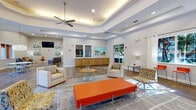 Located at 2450 Lake Debra Dr in Orlando, Florida, this single-floor community facility presents a well-appointed, open-concept interior designed for comfort and social interaction. The space is characterized by striking tray and coffered ceilings fitted with recessed lighting and ceiling fans, creating an airy, elevated atmosphere throughout.

The main lounge area is expansive and thoughtfully furnished with neutral-toned sofas, accent chairs, and bold orange ottomans and accent pieces that inject vibrant energy into the design. Rich wood-look flooring runs throughout, grounded by area rugs that define distinct seating zones. Colorful artwork adorns the walls, complementing the contemporary color palette.

Large windows dressed with white curtains and orange Roman shades allow generous natural light to pour in, offering views of the surrounding landscaped grounds. High-top counter seating with orange barstools lines one section of the space, providing a casual work or gathering area alongside warm wood cabinetry.

A secondary lounge nook features plush gray seating, a mounted flat-screen television, and coordinating mosaic-style wall art, creating an intimate retreat within the larger space. Throughout, the design balances open, communal gathering areas with cozy, defined pockets — making this a versatile and welcoming facility for residents and visitors alike.