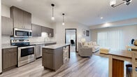 This single-floor unit showcases a thoughtfully designed open-concept layout that seamlessly connects the kitchen, dining, and living areas. The kitchen is appointed with dark espresso cabinetry, granite countertops, and a full suite of stainless steel appliances, including a refrigerator, range, and built-in microwave. A central island with seating provides both additional workspace and a casual dining option, while pendant lighting adds a contemporary touch overhead.

The living area is bright and welcoming, featuring wood-look plank flooring that extends throughout the main living spaces. A large sliding glass door with vertical blinds allows abundant natural light and provides access to an outdoor area. A built-in media niche offers a dedicated space for entertainment.

The bedroom is carpeted for comfort and includes a ceiling fan, wall-mounted TV, and a window with blinds that filters natural light. A dedicated den or office space, also carpeted, features a window and ample room for a desk and shelving unit, making it well-suited for remote work or study.

The bathroom continues the cohesive design aesthetic with matching dark cabinetry, a granite-topped vanity, a well-lit mirror, and wood-look tile flooring. A frosted glass door off the kitchen provides access to a laundry area, completing this functional and modern unit.