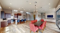 Located on the sixth floor in New Orleans, this clubroom is a well-appointed communal space designed for gatherings and relaxation. The open-plan layout seamlessly connects three distinct zones — a fully equipped kitchen, a formal dining area, and a comfortable lounge — all unified by light wood-look flooring throughout.

The kitchen is a standout feature, outfitted with rich dark-wood cabinetry, stainless steel appliances including a full-size refrigerator and range, a wall-mounted range hood, and a large white-topped island with blue upholstered bar seating. Clerestory windows above draw in additional natural light, enhancing the airy feel of the space.

The dining area accommodates a generous dark wood table surrounded by bold red upholstered chairs, anchored beneath a contemporary chandelier. Floor-to-ceiling black-framed glass doors open directly onto an outdoor terrace, flooding the space with natural light and providing access to exterior views.

The lounge area features a spacious sectional sofa paired with brown leather armchairs, centered around a textured white brick accent wall with a mounted flat-screen television. Roller shades along large windows offer flexible light control. Recessed lighting throughout maintains a clean, modern ambiance across the entire space.