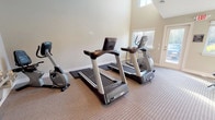 This fitness center offers a well-rounded workout environment within a single open space designed to accommodate a variety of exercise routines. The room features soaring vaulted ceilings that create an open, airy atmosphere, complemented by multiple windows that flood the space with natural light and provide views of the surrounding outdoor greenery. Carpeted flooring runs throughout, lending a clean and maintained appearance.

The cardio zone is equipped with multiple treadmills and an elliptical trainer, as well as a recumbent stationary bike, catering to a range of low- and high-impact aerobic workouts. A dedicated strength and functional training area includes a full rack of dumbbells in varying weights, a set of medicine balls on a vertical stand, and an adjustable workout bench, providing options for both resistance training and dynamic exercises. A wall-mounted flat-screen television is positioned for easy viewing during workouts.

Additional amenities include a wall-mounted water fountain for convenient hydration and access to restroom facilities on-site. A glass-paneled exterior door provides direct outdoor access. The layout ensures ample floor space between equipment, allowing for comfortable movement and stretching. Overall, the fitness center is a practical and well-lit space suited for a comprehensive daily workout routine.