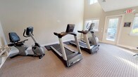 This fitness center offers a well-rounded workout environment within a single open space designed to accommodate a variety of exercise routines. The room features soaring vaulted ceilings that create an open, airy atmosphere, complemented by multiple windows that flood the space with natural light and provide views of the surrounding outdoor greenery. Carpeted flooring runs throughout, lending a clean and maintained appearance.

The cardio zone is equipped with multiple treadmills and an elliptical trainer, as well as a recumbent stationary bike, catering to a range of low- and high-impact aerobic workouts. A dedicated strength and functional training area includes a full rack of dumbbells in varying weights, a set of medicine balls on a vertical stand, and an adjustable workout bench, providing options for both resistance training and dynamic exercises. A wall-mounted flat-screen television is positioned for easy viewing during workouts.

Additional amenities include a wall-mounted water fountain for convenient hydration and access to restroom facilities on-site. A glass-paneled exterior door provides direct outdoor access. The layout ensures ample floor space between equipment, allowing for comfortable movement and stretching. Overall, the fitness center is a practical and well-lit space suited for a comprehensive daily workout routine.