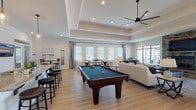 Located at 75 Siskin Cir in Hampstead, NC, this single-story community clubhouse offers a thoughtfully designed gathering space with a variety of amenities. The main lounge area is an expansive, open room anchored by a floor-to-ceiling stacked stone fireplace with a mounted flat-screen TV. Tray ceilings with crown molding add architectural elegance, while large windows and glass doors bathe the space in natural light and offer views of the outdoors. Light hardwood flooring runs throughout, unifying each area with a clean, coastal aesthetic. A ceiling fan overhead provides both comfort and style.

The games and social area features a full-sized billiards table, high-top bar seating, a casual dining table, and ample lounge furniture — all arranged to encourage relaxed gatherings. Adjacent to this is a well-appointed kitchen with dark cabinetry, granite countertops, white subway tile backsplash, stainless steel appliances including a full-sized refrigerator, a coffee station, and pendant lighting.

A dedicated business center provides multiple computer workstations with granite desk surfaces, a printer, and natural light from a nearby window. A separate fitness room is visible through glass walls and is equipped with cardio and strength training equipment. The overall ambiance is bright, modern, and welcoming — ideal for community interaction and recreation.