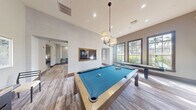 Located at 4600 Portofino Way in West Palm Beach, Florida, this well-appointed community clubhouse offers a thoughtfully designed single-level layout encompassing a generous amount of social and recreational space. The interior is finished with wood-look plank flooring, neutral gray walls, and rich dark wood baseboards, creating a refined coastal aesthetic throughout.

The main living area serves as the social heart of the space, featuring a large, plush sectional sofa in cream upholstery, a sculptural round coffee table, and elegant pendant lighting. Multiple tall windows frame lush outdoor greenery, flooding the room with natural light. Coastal-inspired artwork adorns the walls, reinforcing the relaxed Florida atmosphere.

An adjacent dining nook is defined by a built-in banquette with blue upholstery set against a textured botanical-patterned accent wall, illuminated by stylish pendant lights overhead. A second living area doubles as a dedicated game room, anchored by a contemporary pool table with blue felt beneath a multi-light chandelier. A full-length shuffleboard table runs alongside large windows, and a large mirror amplifies the sense of space. A wall-mounted flat-screen TV adds to the entertainment offerings. The layout connects seamlessly between zones, making this clubhouse an ideal gathering destination for recreation and relaxation alike.