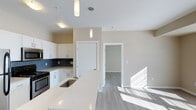 This single-floor space showcases a thoughtfully designed open-concept layout that seamlessly connects the kitchen and living areas. The kitchen is a standout feature, equipped with crisp white cabinetry, a dark tile backsplash, and stainless steel appliances. A generously sized island with a light quartz countertop runs along the center, incorporating a stainless steel sink and providing ample workspace and casual dining potential. Two elegant pendant lights hang above the island, adding a modern touch to the space.

The flooring throughout is a light-toned laminate or vinyl plank, lending a clean, contemporary feel while enhancing the sense of openness. Neutral beige walls complement the white trim and doors, creating a warm yet understated palette. Natural light streams into the living area from a nearby window, brightening the space considerably.

A doorway visible from the kitchen leads to an additional room, suggesting a well-connected floor plan with accessible private spaces. The smooth white ceilings and recessed lighting further contribute to the airy, modern ambiance that defines this well-appointed unit.