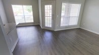 This single-floor space offers a clean, modern aesthetic defined by key design choices that maximize both functionality and visual appeal. The main living area is finished with wide-plank vinyl flooring in a warm gray-brown tone, providing a durable yet stylish foundation that ties the space together seamlessly.

Natural light is a defining characteristic of this space. Multiple large windows dressed with horizontal blinds line the walls, allowing generous amounts of daylight to pour in throughout the day. A central glass-paneled entry door further enhances this brightness, creating an open and welcoming atmosphere while offering a view of the surrounding greenery outside.

The walls are finished in a soft neutral gray, complementing the flooring and contributing to a calm, contemporary ambiance. Clean white trim and baseboards provide crisp contrast throughout, adding a polished, well-maintained feel to the interior.

The layout is open and unobstructed, allowing for flexible furniture arrangement and a sense of spaciousness. The overall design reflects a practical yet attractive aesthetic, well-suited for comfortable everyday living. The combination of natural light, neutral tones, and modern finishes gives this space a fresh, move-in-ready character that is both inviting and functional.