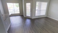 This single-floor space offers a clean, modern aesthetic defined by key design choices that maximize both functionality and visual appeal. The main living area is finished with wide-plank vinyl flooring in a warm gray-brown tone, providing a durable yet stylish foundation that ties the space together seamlessly.

Natural light is a defining characteristic of this space. Multiple large windows dressed with horizontal blinds line the walls, allowing generous amounts of daylight to pour in throughout the day. A central glass-paneled entry door further enhances this brightness, creating an open and welcoming atmosphere while offering a view of the surrounding greenery outside.

The walls are finished in a soft neutral gray, complementing the flooring and contributing to a calm, contemporary ambiance. Clean white trim and baseboards provide crisp contrast throughout, adding a polished, well-maintained feel to the interior.

The layout is open and unobstructed, allowing for flexible furniture arrangement and a sense of spaciousness. The overall design reflects a practical yet attractive aesthetic, well-suited for comfortable everyday living. The combination of natural light, neutral tones, and modern finishes gives this space a fresh, move-in-ready character that is both inviting and functional.