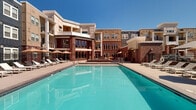 This outdoor amenity space is defined by a striking blend of classic brick and contemporary architectural elements that frame a generously sized resort-style swimming pool at its center. The pool deck is thoughtfully designed, offering an abundance of lounge seating, including rows of chaise lounges arranged along the poolside for sun bathing and relaxation. Shaded umbrella clusters provide respite from direct sunlight, creating comfortable gathering zones throughout the deck.

Adjacent to the main pool, a separate spa or wading feature adds to the leisure options available. A dedicated covered lounge area—finished with white shiplap-style walls and outfitted with modern tan-upholstered armchairs and side tables—provides a sheltered retreat for casual conversation and relaxation.

A secondary courtyard area features additional modern outdoor seating arrangements set against a dramatic red brick facade with tall, multi-paned windows and upper-level balconies. This intimate space is anchored by a raised planter or fire feature, lending a sophisticated, community-focused atmosphere.

The surrounding multi-story building incorporates varied façade materials—brick, painted siding, and neutral stucco—with numerous balconies overlooking the courtyard. Lush greenery borders portions of the property, softening the hardscape and enhancing the overall open-air ambiance of this vibrant outdoor community hub.
