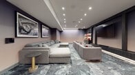 Located at 1000 Schindler Dr in South Amboy, NJ, this single-floor cinema lounge is designed to deliver an immersive, theater-inspired experience. The main screening area is anchored by a large wall-mounted display framed by dark, richly paneled built-ins with integrated shelving, displaying curated décor pieces and accent lighting. Generous sectional sofas in soft grey upholstery, accented with bold red and charcoal throw pillows, are arranged to maximize viewing comfort across the space. A patterned carpet in cool grey and blue tones runs throughout, adding texture and visual depth. The walls are adorned with large-scale, framed black-and-white cinema posters—including classics such as Breakfast at Tiffany's, Rocky, Annie Hall, and War of the Worlds—reinforcing the space's cinematic identity. An adjoining lounge and refreshment area features light hardwood flooring, a sleek marble-topped island with bar seating, and additional cabinetry for storage. Modern pendant lighting and recessed ceiling fixtures illuminate this zone warmly. A seating nook with vibrant red accent chairs and a small coffee table provides an informal gathering spot near the entrance. Recessed and track lighting throughout creates a polished, contemporary ambiance suited for both casual gatherings and cinematic events.