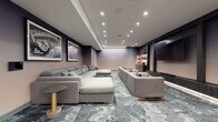 Located at 1000 Schindler Dr in South Amboy, NJ, this single-floor cinema lounge is designed to deliver an immersive, theater-inspired experience. The main screening area is anchored by a large wall-mounted display framed by dark, richly paneled built-ins with integrated shelving, displaying curated décor pieces and accent lighting. Generous sectional sofas in soft grey upholstery, accented with bold red and charcoal throw pillows, are arranged to maximize viewing comfort across the space. A patterned carpet in cool grey and blue tones runs throughout, adding texture and visual depth. The walls are adorned with large-scale, framed black-and-white cinema posters—including classics such as Breakfast at Tiffany's, Rocky, Annie Hall, and War of the Worlds—reinforcing the space's cinematic identity. An adjoining lounge and refreshment area features light hardwood flooring, a sleek marble-topped island with bar seating, and additional cabinetry for storage. Modern pendant lighting and recessed ceiling fixtures illuminate this zone warmly. A seating nook with vibrant red accent chairs and a small coffee table provides an informal gathering spot near the entrance. Recessed and track lighting throughout creates a polished, contemporary ambiance suited for both casual gatherings and cinematic events.