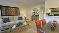 This single-floor residence offers a comfortable and cohesive living environment with a thoughtfully arranged open-concept layout. The living area is anchored by plush wall-to-wall carpeting that lends a warm, inviting feel throughout the space. Soft, neutral-toned walls create a calm backdrop, complemented by recessed lighting that adds a gentle brightness to the interior.

The living room provides generous space for comfortable seating and features a glass-top coffee table and coordinating side table, contributing to a polished yet approachable aesthetic. A large framed artwork accents the main wall, adding a touch of character to the room.

The dining area sits adjacent to the living space and is defined by a rich, dark dining table paired with tufted leather-style chairs in a warm reddish-orange tone, offering a bold contrast to the otherwise understated palette.

Flowing naturally from the living and dining areas is a dedicated cabinet and shelving zone, featuring wood-toned cabinetry with display shelving above — ideal for books, décor, and personal items. Track lighting above the shelving highlights the display area, adding both function and ambiance.

Overall, the space presents a harmonious blend of comfort and style, making it well-suited for everyday living and casual entertaining.