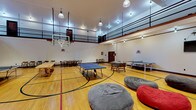 Located at 520 4th Street in Cheney, Washington, this recreational gymnasium is a versatile, single-floor facility designed to accommodate a wide range of activities and events. The centerpiece of the space is a full-sized hardwood basketball court, complete with painted court lines, a center-circle logo, and retractable basketball hoops at each end. The soaring ceiling height allows for unobstructed play and contributes to the open, athletic atmosphere of the room.

Encircling the court on an upper level is a mezzanine balcony with metal railings, providing an elevated vantage point for spectators or additional overflow use. Below, portable wooden bleachers are positioned along one wall, offering tiered seating for audiences during organized events or games. Pendant-style industrial lighting hangs from the ceiling, ensuring the court is well-lit throughout.

The gymnasium is currently outfitted with multiple recreational amenities, including ping-pong tables, a foosball table, a large wooden gathering table, and bean bag seating, making it suitable for social or multi-use gatherings. A wall-mounted flat-screen television is installed on one side of the court. Vintage wooden chairs are arranged along the perimeter walls, and dark wood baseboards complement the clean white walls, giving the space a structured yet welcoming character.