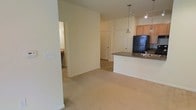 This single-floor residence features an open-concept design that integrates the living area and kitchen into a cohesive, functional space. The living area is finished with wall-to-wall neutral carpeting, lending a warm and understated aesthetic. Light-colored walls throughout enhance the sense of openness and brightness.

The kitchen is positioned along one side of the main living space and is defined by a peninsula-style counter with a dark granite or stone-look countertop, providing both workspace and a casual bar-seating area. A pendant light hangs above the peninsula, adding a focused decorative element. The kitchen is equipped with wooden cabinetry in a warm tone, a black refrigerator, and a stainless steel sink. Track lighting above the kitchen ensures ample task illumination.

Adjacent to the living area, a hallway leads to additional rooms, including what appears to be a closet with paneled double doors. A glimpse into a secondary room reveals tile flooring and additional cabinetry, suggesting a bathroom or utility space. The overall layout is efficient and well-organized, offering a clean, move-in-ready environment suited for everyday living.