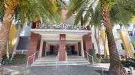 Boca City Walk is a residential community that makes a strong first impression through its well-designed exterior entrance. Building 33 serves as a focal point, featuring a covered entryway with clean, contemporary architectural lines highlighted by bold red and neutral paneling. The entrance is elevated by a short flight of steps with accessible ramps on either side, ensuring ease of access for all residents and visitors. Large, bold metallic lettering spelling "Boca City Walk" crowns the façade, lending the property a distinctive identity. Flanking the entrance, tall mature palm trees add a tropical, Florida-inspired ambiance that complements the warm exterior color palette. Blue decorative planters are positioned within the entryway corridor, adding subtle touches of color to the passage. A clearly marked directional sign points visitors toward the on-site leasing center, indicating a well-organized and professionally managed community. The architecture of the building reflects a modern, mixed-use residential aesthetic, with additional building structures visible in the background showcasing varied architectural accents. The overall design presents a vibrant, welcoming community environment that combines functional accessibility with contemporary curb appeal.