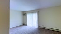 This single-floor space offers a clean and functional layout designed for comfortable everyday living. The main living area is finished with wall-to-wall carpeting that adds warmth underfoot and contributes to a quiet, cozy atmosphere. A large sliding glass door serves as the focal point of the room, dressed with vertical blinds that allow for adjustable light control and privacy. Natural light filters through the door, brightening the neutral, light-toned walls and giving the space an open, airy feel. A wall-mounted air conditioning unit is installed for efficient climate control during warmer months, while continuous baseboard heating runs along the perimeter of the room, ensuring consistent warmth during cooler seasons. The smooth white ceiling and simple, unadorned walls create a versatile backdrop well-suited to a variety of interior styles. The overall ambiance is practical and understated, offering a blank canvas that can be personalized to suit individual tastes. The combination of natural light, dual-climate systems, and soft carpeting makes this a well-equipped and livable space.