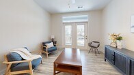 Located in Knightdale, NC, this well-appointed one-bedroom, one-bathroom unit offers a thoughtfully designed single-floor layout that maximizes functionality and comfort. The living room is bright and welcoming, featuring light wood-look flooring, a ceiling fan, and French doors with transom windows that open onto a private balcony, filling the space with natural light and outdoor views. Adjacent to the living area, the dining room is distinguished by a built-in wall of open shelving—ideal for display or storage—and an elegant pendant chandelier overhead. The kitchen is a standout feature, equipped with gray shaker-style cabinetry, granite countertops, a subway tile backsplash, stainless steel appliances including a refrigerator, range, microwave, and dishwasher, plus a peninsula for additional prep space. The generously sized primary bedroom is carpeted for comfort, features a window with natural light, and connects to a walk-in closet outfitted with wire shelving systems. The bathroom is finished with wood-look flooring, a gray vanity with granite countertops, a framed mirror, and stylish lighting. Clean neutral tones and consistent finishes throughout create a cohesive, modern aesthetic.