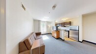 Located at 20 International Place in Tewksbury, MA, this two-bedroom, two-bathroom suite offers a practical and well-appointed single-floor layout. The open-concept living and kitchen area serves as the heart of the suite, where natural light streams through multiple windows framed by sheer white curtains. The living area features plush carpeting and a comfortable sofa, while a half-wall peninsula with granite countertops provides a natural division from the kitchen. The kitchen is equipped with dark espresso cabinetry, stainless steel appliances including a full-size refrigerator, gas range, and a built-in microwave hood, all set on hardwood-style flooring. Both bedrooms are carpeted and feature warm wood-paneled headboards, ample natural light from windows with outdoor views, and dedicated closet space. One bedroom includes a work desk, offering a functional workspace. Each of the two bathrooms features hardwood-style flooring, granite-topped vanities with dark cabinetry, and a shower-tub combination with a clean white curtain. The overall ambiance is bright, neutral, and functional, with a consistent warm-toned palette throughout.