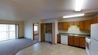 This single-floor space offers a functional and straightforward layout designed for comfortable everyday living. The kitchen is equipped with warm oak cabinetry, dark laminate countertops, a double-basin sink, and a white dishwasher, all illuminated by under-cabinet and overhead fluorescent lighting. The vinyl tile flooring in the kitchen and main area provides durability and easy maintenance. Adjacent to the kitchen is a dedicated laundry closet, housing a full-sized washer and dryer set behind bifold doors, offering the convenience of in-unit laundry. The open living area flows naturally from the kitchen and transitions to plush carpeting, creating a cozy and distinct lounging space. A baseboard heater along the wall ensures efficient climate control throughout the colder months. A doorway leads to a bright bedroom, which benefits from natural light through a window and continues the carpeted flooring. The overall design is practical and well-organized, with neutral tones, light-colored walls, and textured ceilings contributing to a clean, understated ambiance throughout the space.