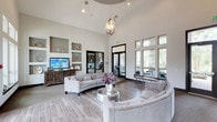 This single-floor space makes an immediate impression with its dramatic vaulted ceilings that lend an airy, expansive feel to the interior. A striking sculptural chandelier serves as a focal point overhead, adding a touch of refined elegance to the open layout. The main lounge area is furnished with plush, tufted sofas and a curved sectional arranged around a sleek metallic accent table, creating an inviting setting for conversation and relaxation. Light wood-toned flooring runs throughout, complementing the soft, neutral color palette of warm whites and greiges that defines the space. Built-in shelving niches are set into the walls, offering both decorative display space and a mounted flat-screen television. Floor-to-ceiling windows along one wall flood the interior with generous natural light while framing views of lush greenery outside. Glass-panel doors provide a seamless visual connection to an adjacent area, enhancing the sense of openness and flow. Black window and door trim provides clean contrast against the bright walls, adding a contemporary architectural detail. Overall, the space balances modern sophistication with comfortable, functional design, making it well-suited as a welcoming communal or gathering area.